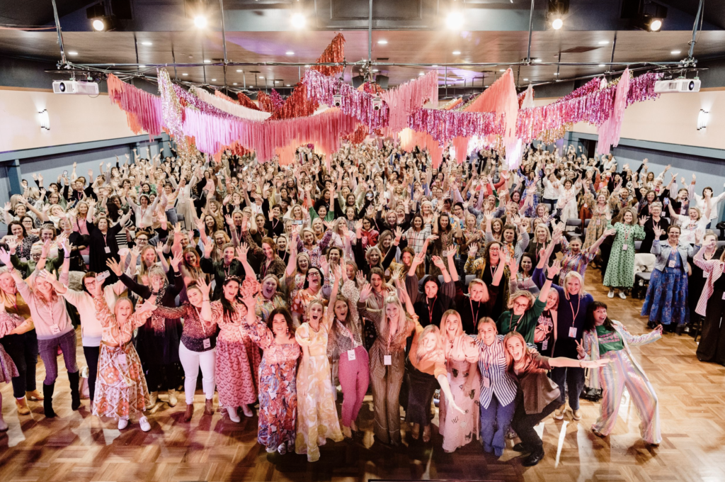 group shot of the huddle event in orange 2022 run by jumbled online with over 700 woman gathered for a conference with pink and orange tinsel decoration hanging overhead.
