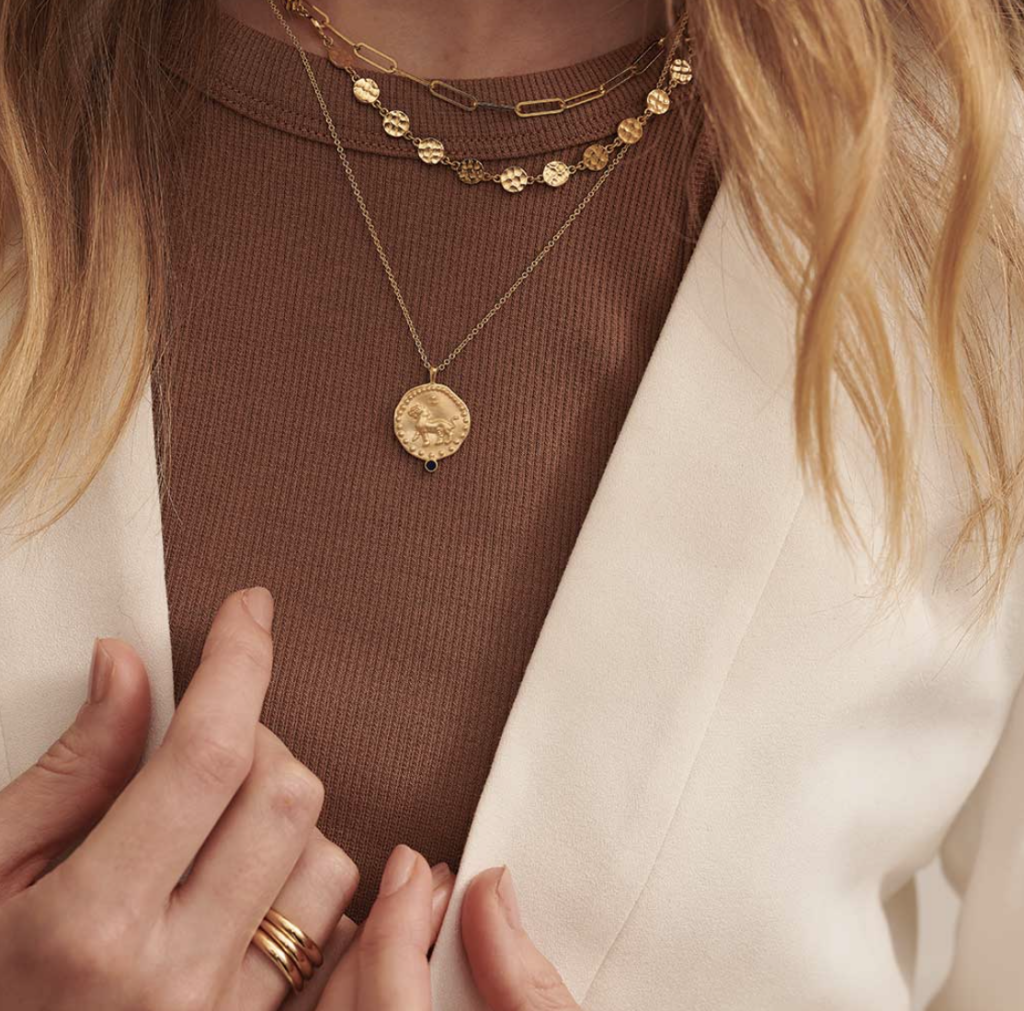 lifestyle shot of murkani gold necklaces layered  with a pendant with lion emblem, and two chokers woman wearing brown ribbed shirt.