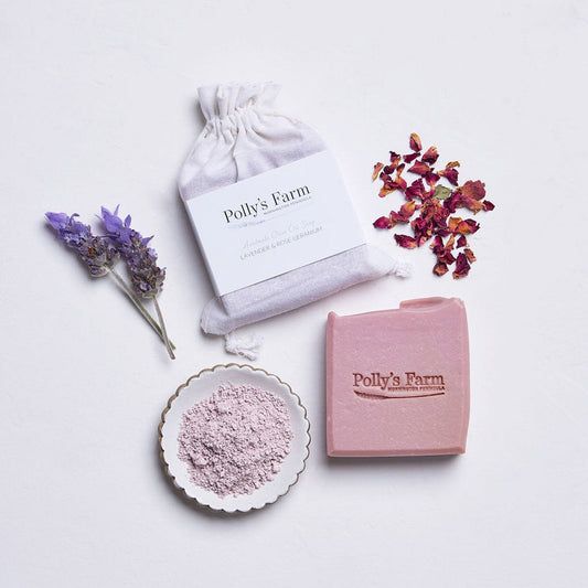 Flat lay of natural skincare products from Polly’s Farm, including a pink bar soap, a small cloth pouch labeled "Lavender & Rose Calming," a bowl of pink powder, dried rose petals, and sprigs of lavender, all arranged on a white background for a clean and calming aesthetic.