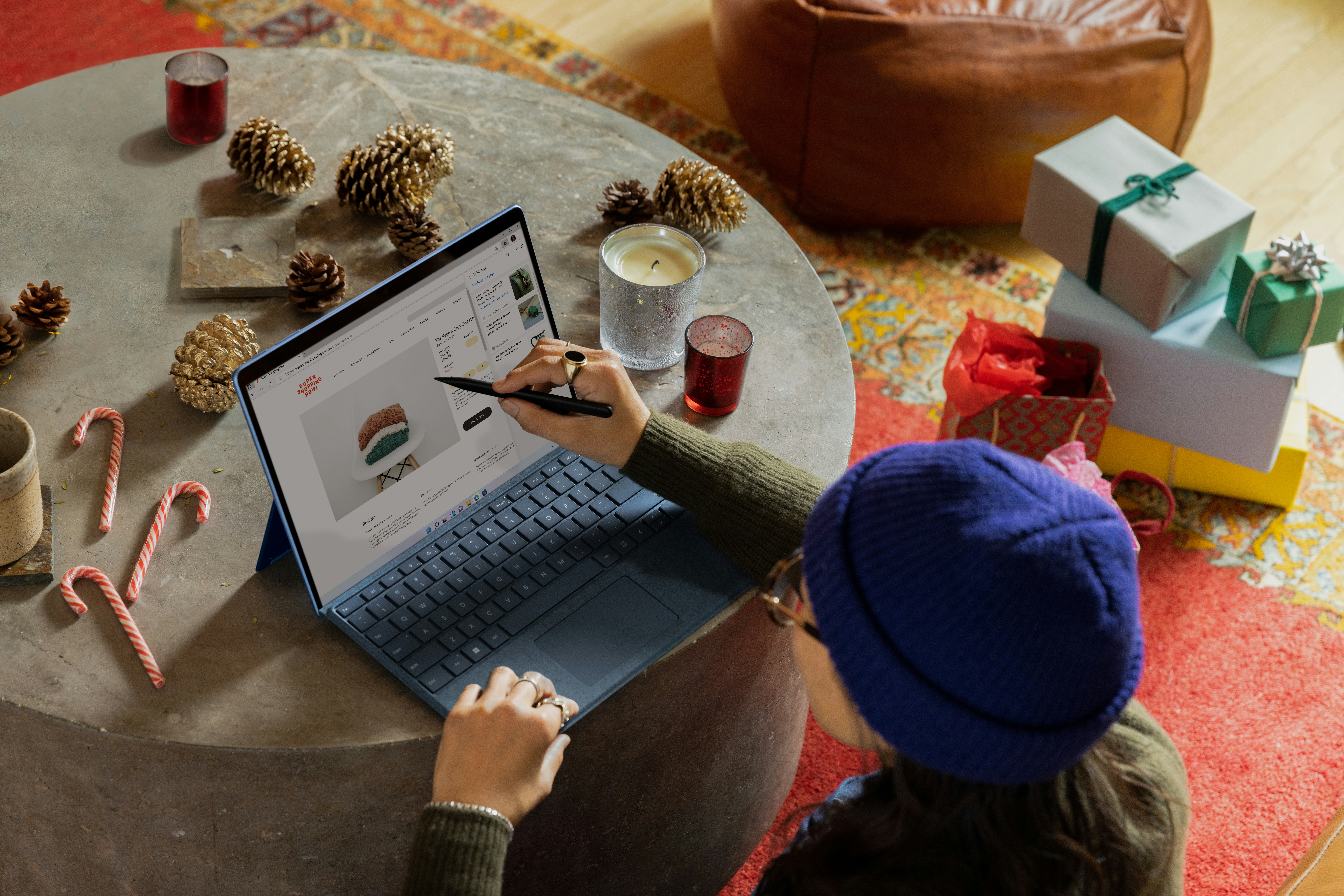 A woman wearing a blue beanie and glasses is sitting at a round coffee table, using a stylus to browse an online store on a blue laptop. The table is decorated with pinecones, candles, and candy canes, and wrapped gifts are visible in the background, creating a festive holiday atmosphere.