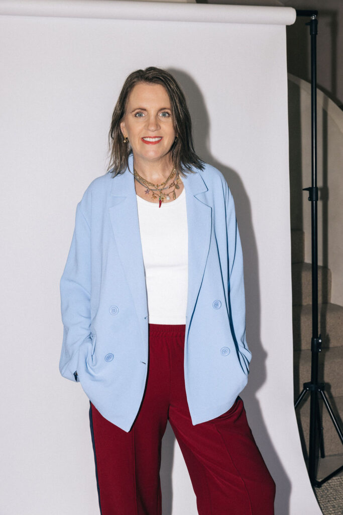 melissa robbins business coach for ecommerce and wholesale strategy standing in front of white photo wall with pale blue blazer on and red pants and white top.